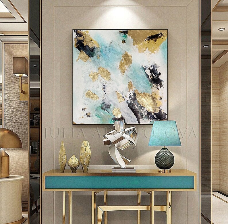 May include: Abstract painting with gold leaf accents on a white canvas, displayed above a teal console table with gold legs. The table is decorated with a silver metal sculpture and three gold ceramic vases.