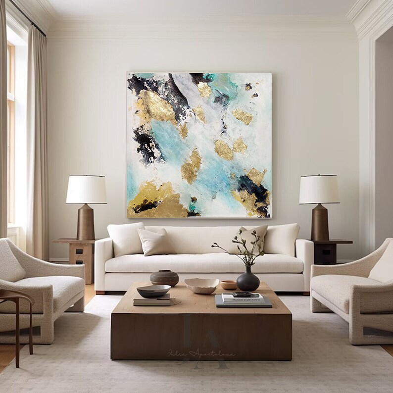A modern living room featuring a large abstract painting with turquoise, gold, black, and white colors on the wall. The room has a light-colored sofa with beige cushions, two matching armchairs, table lamps, and a wooden coffee table in the center.