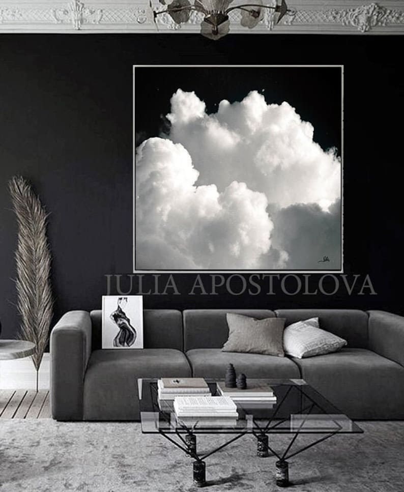 A large, framed black and white cloudscape art piece hangs above a gray sofa. The artwork features a dramatic depiction of fluffy white clouds against a dark sky. The room is decorated in shades of gray and black, with a modern aesthetic.