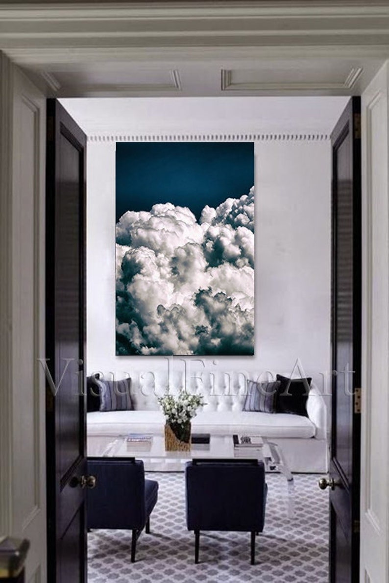May include: A white couch with gray and black throw pillows sits in a room with a large canvas print of white clouds against a blue sky. Two dark blue chairs are in front of the couch, and a glass coffee table sits between them.