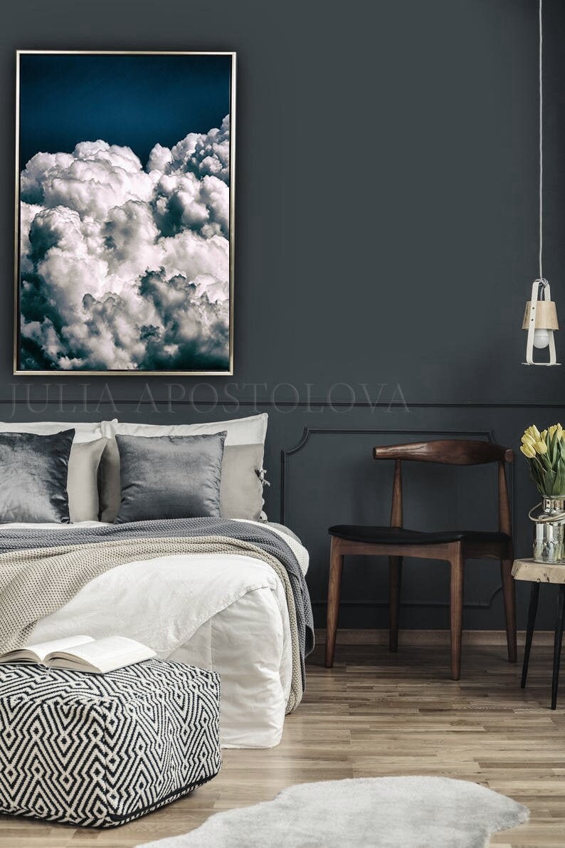 May include: A bedroom interior with a bed, a chair, a footstool, and a framed photograph of a cloudy sky. The walls are painted a dark blue color, and the floor is made of wood.