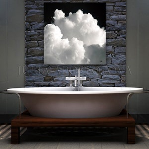 May include: A modern bathroom featuring a white oval bathtub on a dark wood base. Above the tub, a framed artwork depicts a dramatic cloud formation against a black sky. The wall behind the tub is made of gray stone.