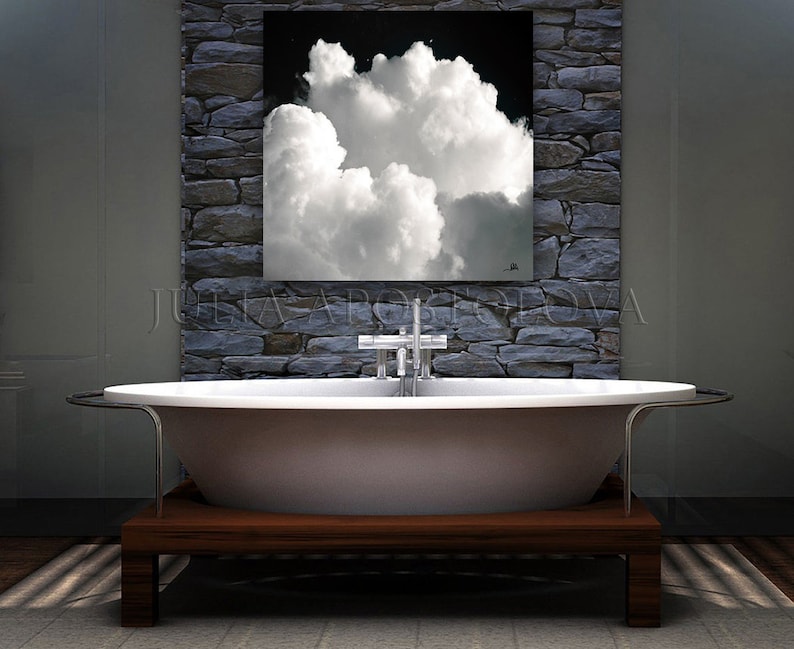 May include: A modern bathroom featuring a white oval bathtub on a dark wood base. Above the tub, a framed artwork depicts a dramatic cloud formation against a black sky. The wall behind the tub is made of gray stone.