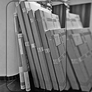 May include: A stack of cardboard boxes and wrapped items, some with "FRAGILE" tape. The items are leaning against a wall, suggesting they are ready for shipping or storage. The image is in black and white.