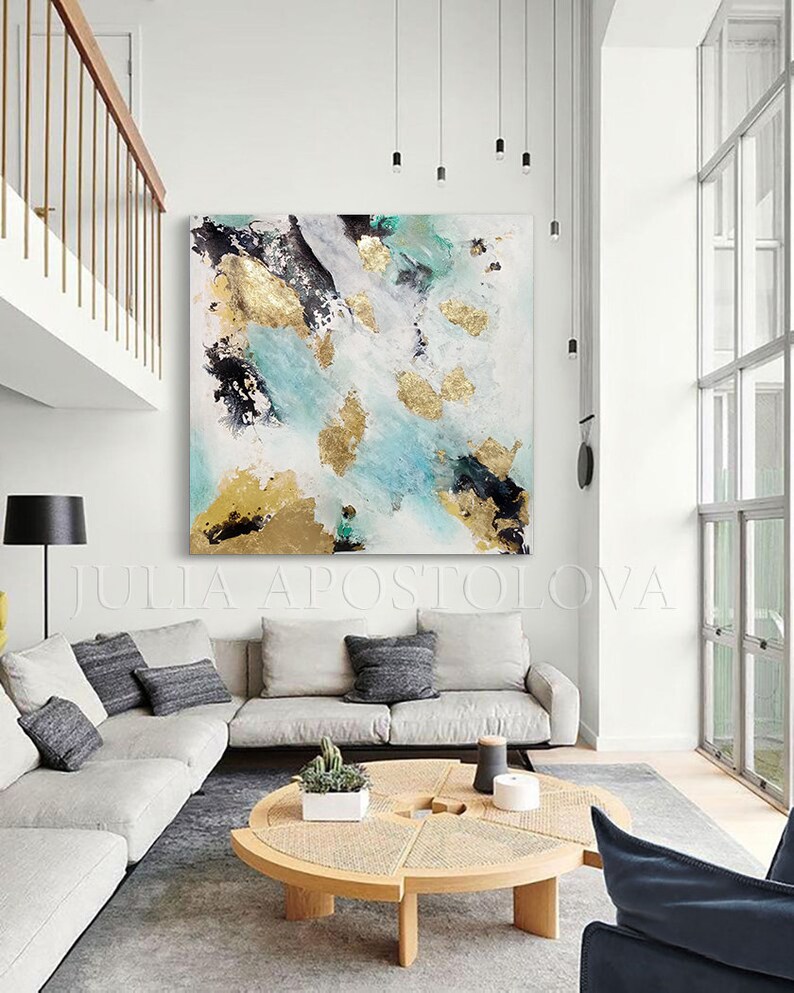 May include: Abstract painting with gold, black, and teal accents on a white canvas. The painting is hanging on a white wall in a modern living room with a white couch, a coffee table, and a black chair.