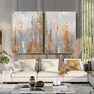 May include: Two framed abstract paintings with gold, gray, and white textures hang above a white sofa. The paintings are rectangular with gold frames. The text "JULIA APOSTOLOVA" is visible below the paintings. The room has a modern design.