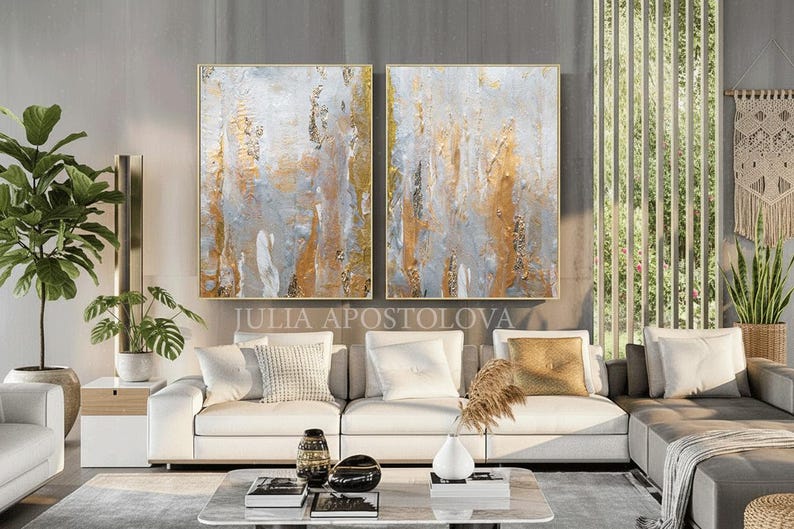 May include: Two framed abstract paintings with gold, gray, and white textures hang above a white sofa. The paintings are rectangular with gold frames. The text "JULIA APOSTOLOVA" is visible below the paintings. The room has a modern design.