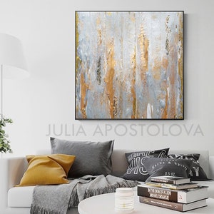 May include: Abstract wall art with a black frame. The artwork features vertical brushstrokes in silver, gold, and gray. The artist's name, JULIA APOSTOLOVA, is printed at the bottom. The painting hangs above a white sofa with gray and yellow pillows.