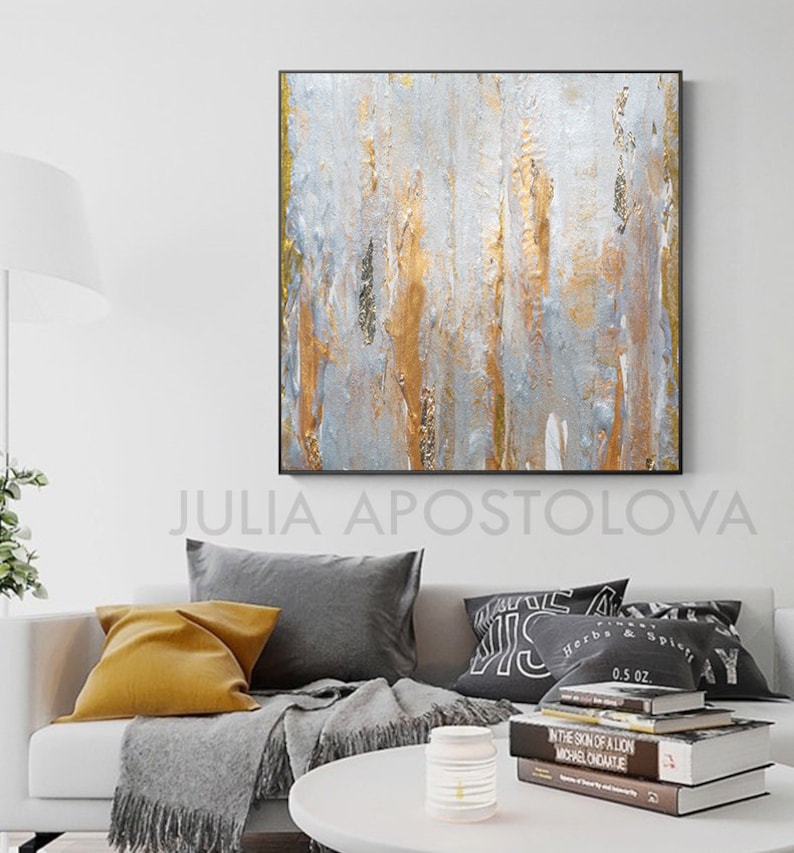 May include: Abstract wall art with a black frame. The artwork features vertical brushstrokes in silver, gold, and gray. The artist's name, JULIA APOSTOLOVA, is printed at the bottom. The painting hangs above a white sofa with gray and yellow pillows.
