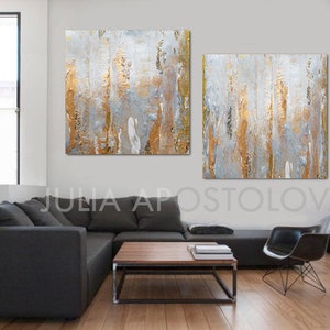 May include: Abstract wall art featuring two square canvases with textured gold, gray, and white vertical strokes. The artwork is displayed in a modern living room setting with a dark gray sectional sofa and a black chair.
