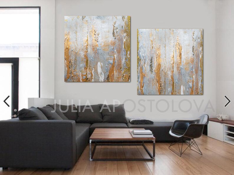 May include: Abstract wall art featuring two square canvases with textured gold, gray, and white vertical strokes. The artwork is displayed in a modern living room setting with a dark gray sectional sofa and a black chair.