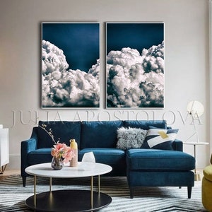 May include: Two framed canvas prints of a blue sky with white fluffy clouds. The prints are hanging on a wall above a blue couch with a white throw pillow and a yellow throw pillow.