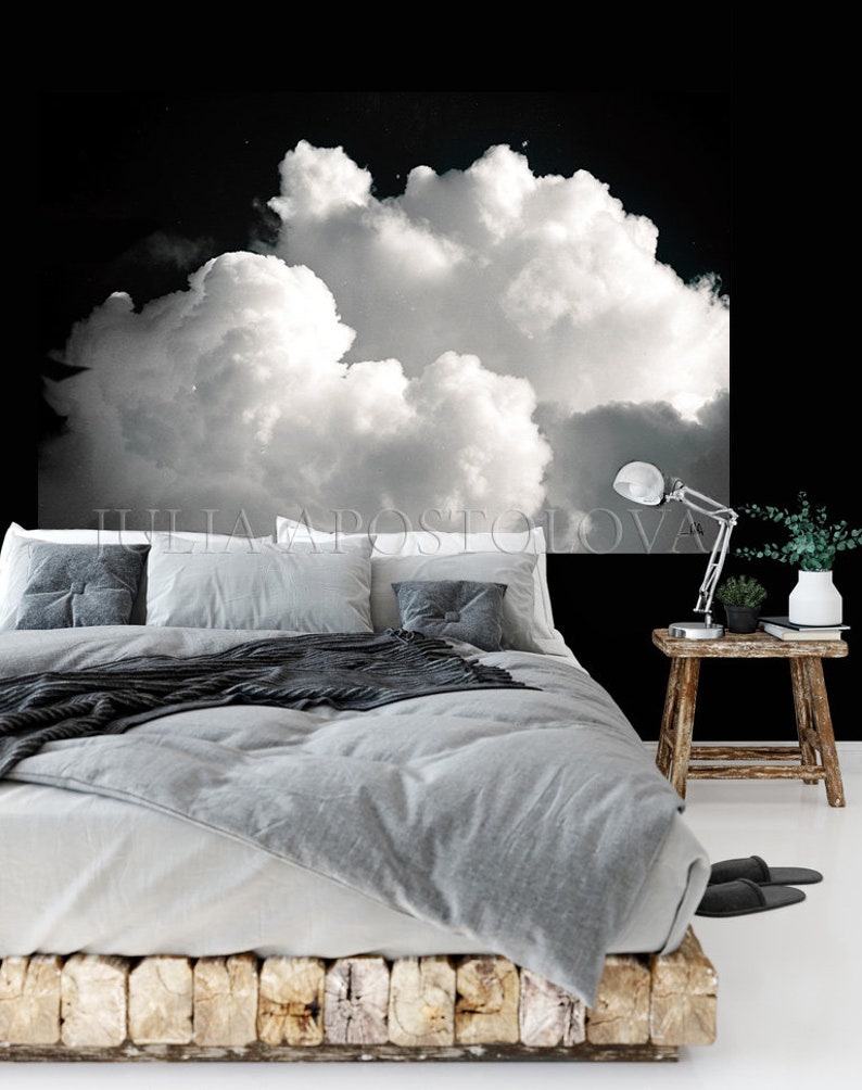 May include: A bedroom scene featuring a bed with gray bedding, pillows, and a dark gray throw. A large black and white cloud mural hangs above the bed. A small wooden side table holds a lamp and plant.