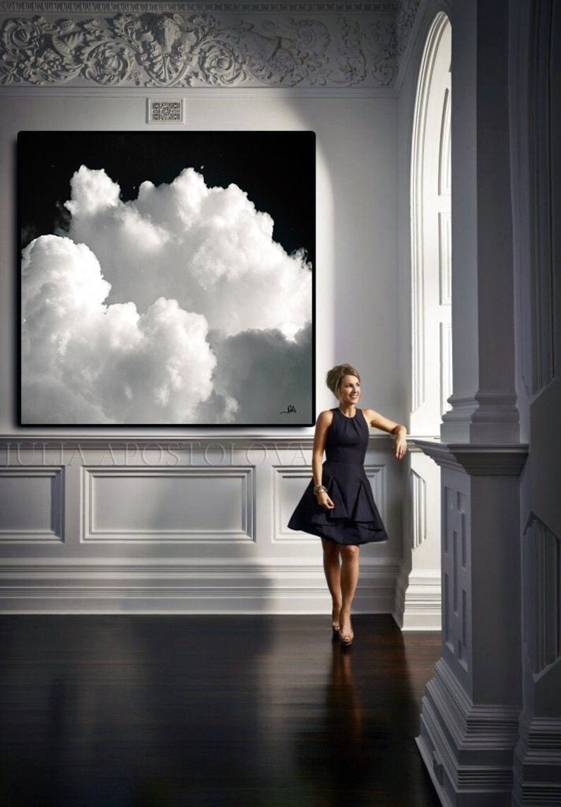 A large black and white cloudscape art piece hangs on a white wall. The artwork features a dramatic depiction of fluffy white clouds against a dark background. A woman in a black dress stands nearby.