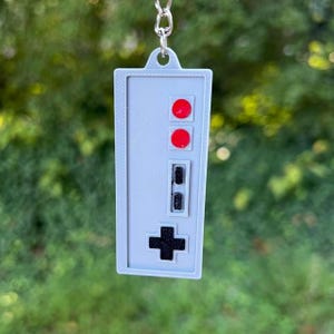 May include: A gray rectangular keychain featuring a classic video game controller design. It has two red circular buttons, a rectangular black button, and a black cross-shaped directional pad. The keychain is suspended from a silver chain.