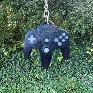 May include: A black plastic Nintendo 64 controller keychain. The controller has a gray faceplate with buttons and a silver analog stick.