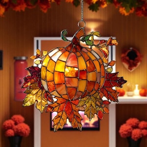 Puede incluir: Una decoración de calabaza de vidriera con un diseño de mosaico en tonos naranja, amarillo y rojo. La calabaza está rodeada de coloridas hojas de otoño en oro, naranja y rojo. Se adjunta una cadena de metal para colgar.