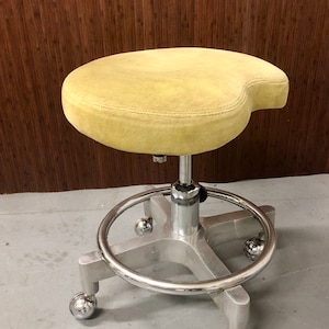 May include: A swivel stool with a kidney-shaped, light yellow upholstered seat. The base is constructed from brushed metal with a circular footrest ring and four legs, each equipped with a small, spherical, polished chrome caster wheel for mobility.
