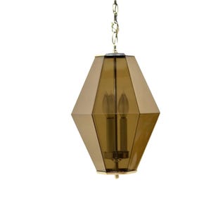 Könnte beinhalten: Eine geometrische Pendelleuchte mit einem goldfarbenen Metallrahmen und einem facettierten, bernsteinfarbenen Glasschirm. Die Leuchte hat zwei kerzenförmige Glühbirnen und hängt an einer goldenen Kette. Das Design ist modern.