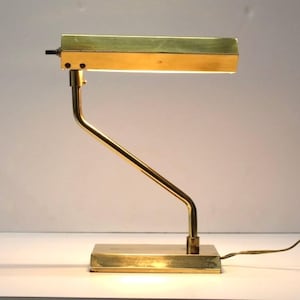 May include: A brass desk lamp with a rectangular shade and a curved arm. The lamp is switched on and illuminating the surface below.