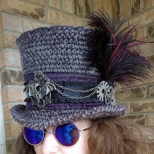 Op de afbeelding: Een grijze en paarse steampunk-stijl hoge hoed met een vleermuis hanger, ketting en tandwiel versieringen. De hoed heeft zwarte en paarse veren en een zwart lint. De persoon draagt blauwe spiegelende zonnebrillen.
