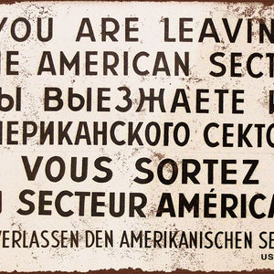 May include: A vintage-style metal sign with a white background and black text. The sign reads "You are leaving the American Sector" in English, Russian, French, and German. The sign also includes the text "US Army".