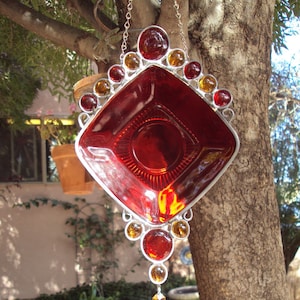 May include: A handcrafted, red glass suncatcher with a diamond-shaped center and a decorative border of amber and red glass beads. The suncatcher is suspended by a silver chain and features a dangling crystal pendant, perfect for garden decor.