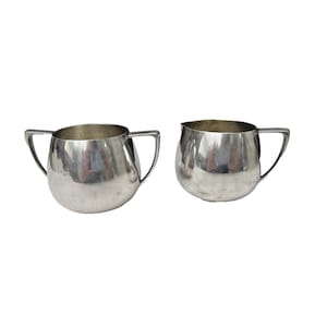 May include: Two silver-colored cream and sugar bowls. Each bowl has a rounded body and a handle. The bowls are made of a shiny, reflective metal. The bowls are set against a white background.