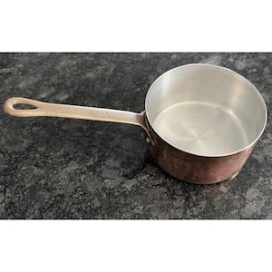 Könnte beinhalten: Ein Kupfer-Kochtopf mit einem langen, bronzefarbenen Griff. Das Innere des Topfes ist silberfarben. Der Topf steht auf einer dunklen, gesprenkelten Oberfläche. Küchenutensilien.