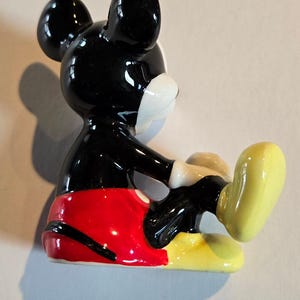 Pu&ograve; includere: Una statuetta in ceramica di Topolino, con testa e orecchie nere lucide, viso bianco e pantaloncini rossi. La figura &egrave; in posa con una gamba estesa, che mostra scarpe gialle e guanti bianchi. Un classico oggetto da collezione Disney.