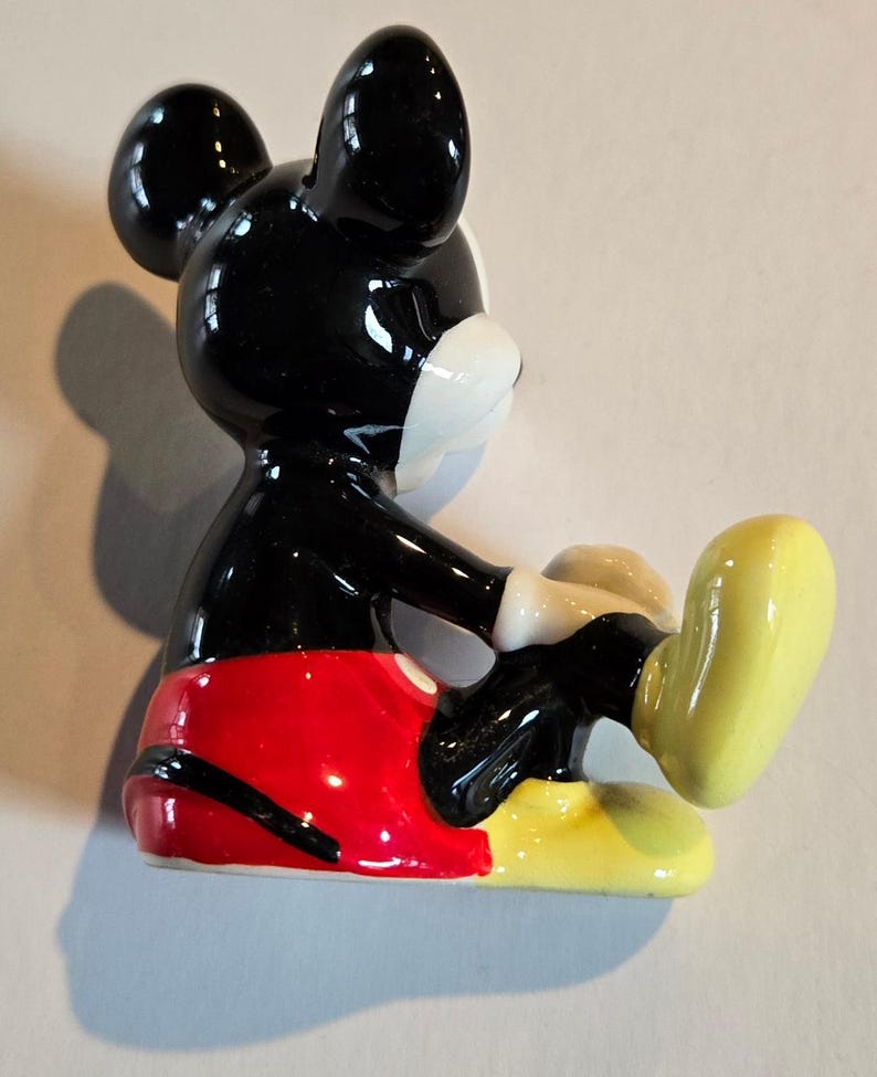 Pu&ograve; includere: Una statuetta in ceramica di Topolino, con testa e orecchie nere lucide, viso bianco e pantaloncini rossi. La figura &egrave; in posa con una gamba estesa, che mostra scarpe gialle e guanti bianchi. Un classico oggetto da collezione Disney.