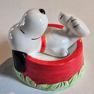 May include: A ceramic Snoopy figurine in his iconic red doghouse. The white Snoopy figure has black ears and a black spot on his nose, with a red collar. The doghouse is red with a green grass base.