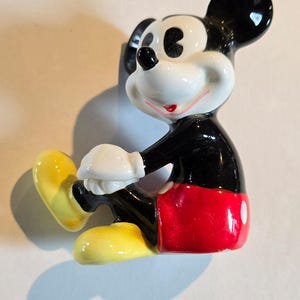 Pu&ograve; includere: Una statuina in ceramica di Topolino in posizione seduta. La figura ha un corpo nero, pantaloncini rossi, scarpe gialle e guanti bianchi. Le orecchie sono nere e il viso bianco con un sorriso rosso.