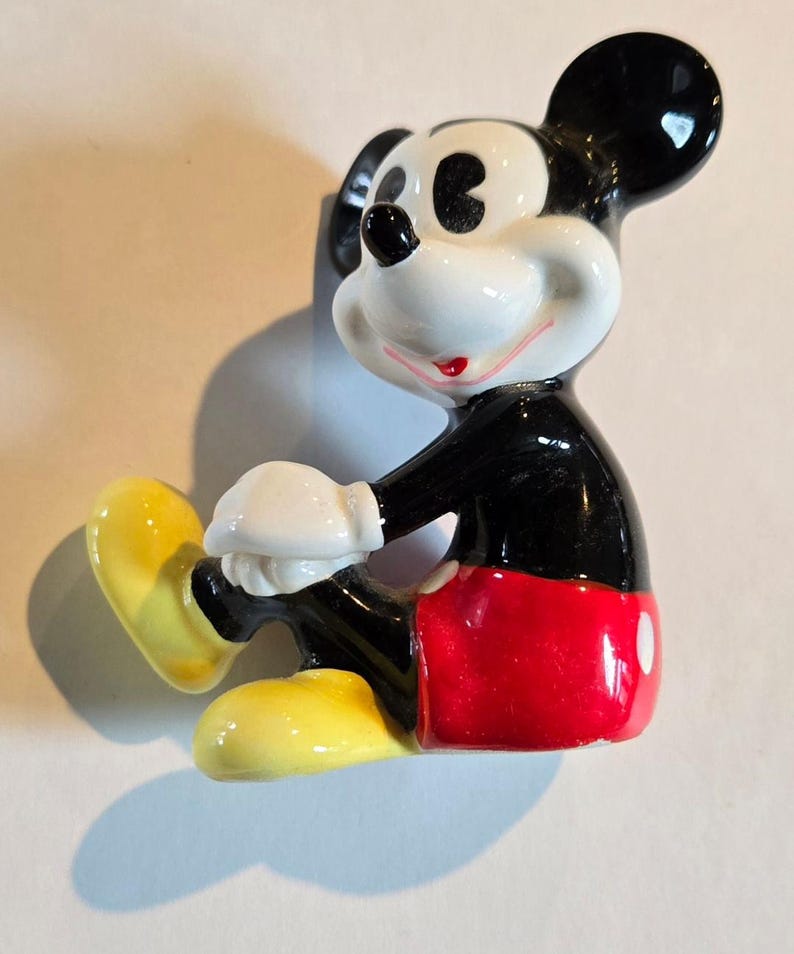 Pu&ograve; includere: Una statuina in ceramica di Topolino in posizione seduta. La figura ha un corpo nero, pantaloncini rossi, scarpe gialle e guanti bianchi. Le orecchie sono nere e il viso bianco con un sorriso rosso.