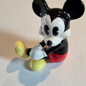 Pu&ograve; includere: Una statuina in ceramica di Topolino in posizione seduta. La figura ha orecchie nere, corpo nero, guanti bianchi e scarpe gialle. Sono visibili anche i pantaloncini rossi e il viso bianco di Topolino. Un classico oggetto da collezione Disney.