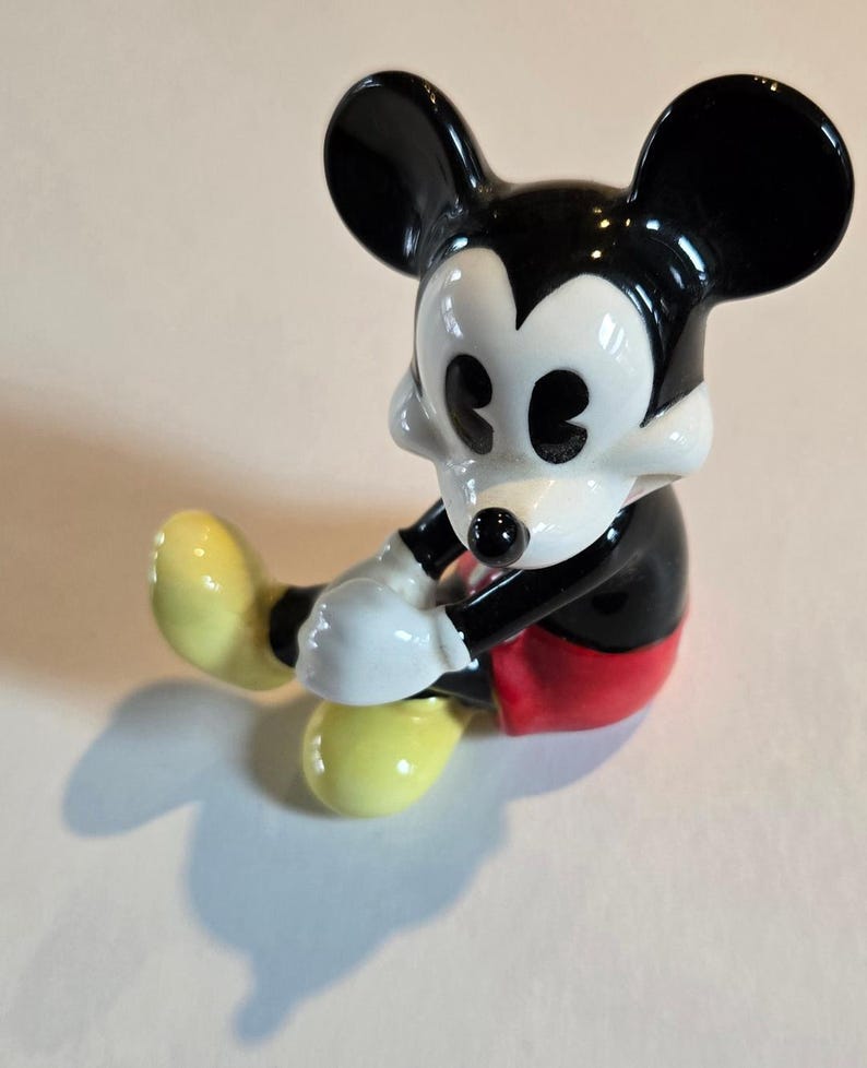 Pu&ograve; includere: Una statuina in ceramica di Topolino in posizione seduta. La figura ha orecchie nere, corpo nero, guanti bianchi e scarpe gialle. Sono visibili anche i pantaloncini rossi e il viso bianco di Topolino. Un classico oggetto da collezione Disney.