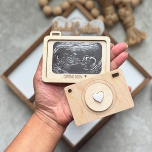 May include: Two light-colored wooden camera-shaped frames. One frame displays a black and white ultrasound image with the text "COMING SOON..." and a small heart. The other frame features a circular lens detail with a small white heart in the center.