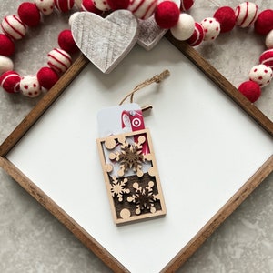 May include: A wooden gift card holder with a cutout design of snowflakes. The holder is decorated with a red and white gift card with a picture of Santa Claus. The holder is sitting on a white surface with a red and white garland.