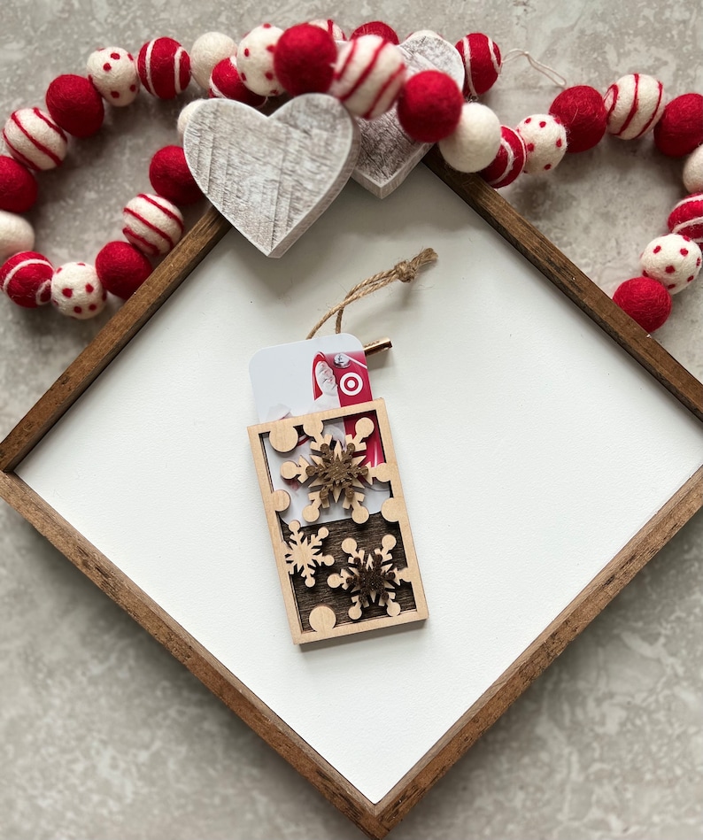 May include: A wooden gift card holder with a cutout design of snowflakes. The holder is decorated with a red and white gift card with a picture of Santa Claus. The holder is sitting on a white surface with a red and white garland.