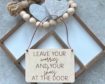 May include: Wooden sign with the text "Leave your worries and your shoes at the door". The sign is hanging from a brown cord and is in front of a white background.