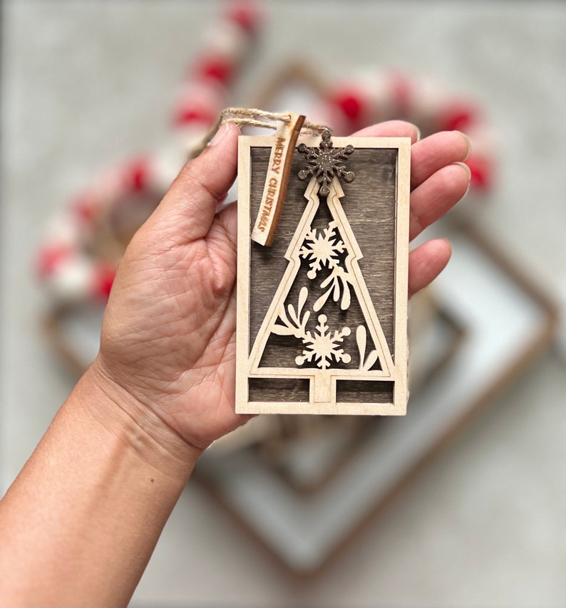 May include: A wooden Christmas tree ornament with a cutout design of snowflakes and leaves. The ornament has a brown woodgrain finish and a small tag that reads "Merry Christmas".