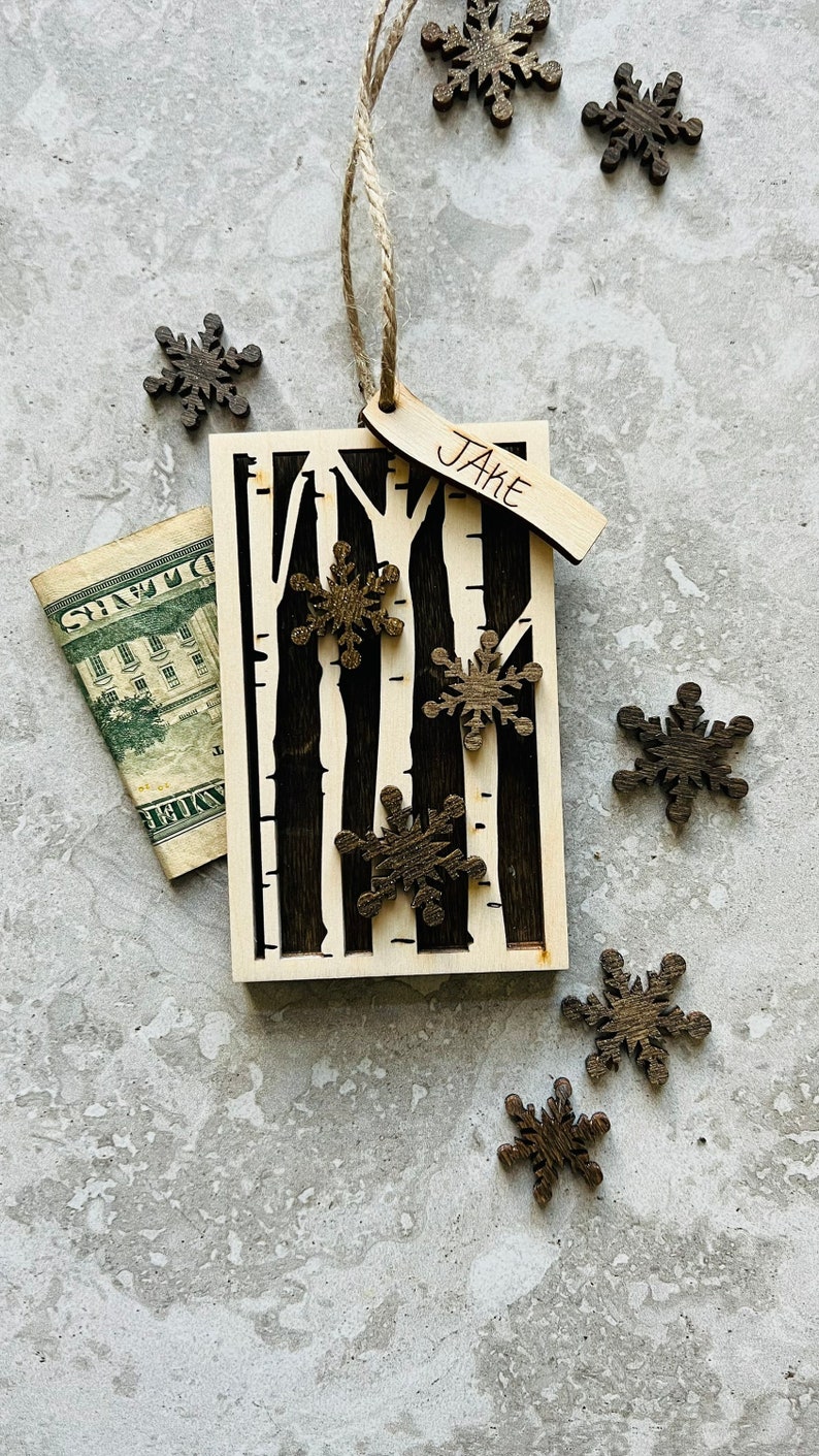 May include: A wooden Christmas ornament with a birch tree design and the name "Jake" written on a tag. The ornament is decorated with small wooden snowflakes.