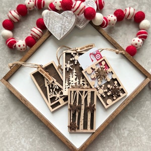 May include: Four wooden Christmas gift tags with different designs. Each tag has a "Merry Christmas" inscription and features a snowflake, tree, or birch tree design. One tag has a gift card slot.