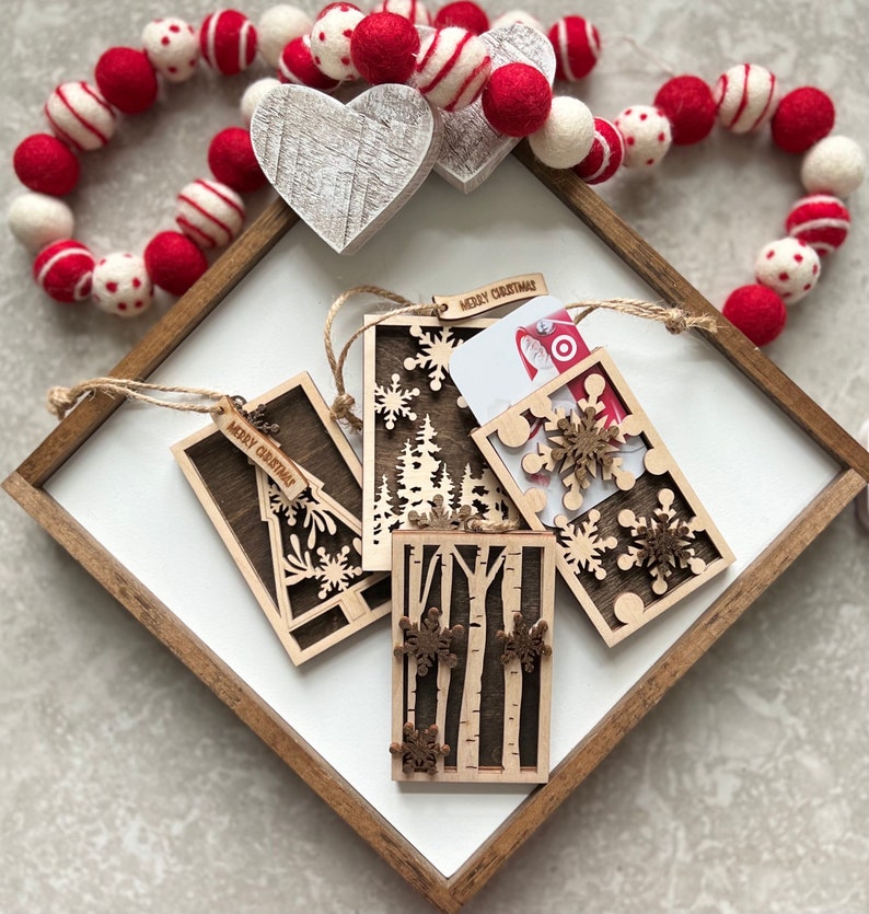 May include: Four wooden Christmas gift tags with different designs. Each tag has a "Merry Christmas" inscription and features a snowflake, tree, or birch tree design. One tag has a gift card slot.