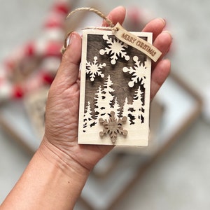 May include: A wooden Christmas ornament with a cutout design of a forest and snowflakes. The ornament is brown and has a small wooden tag that says "Merry Christmas".