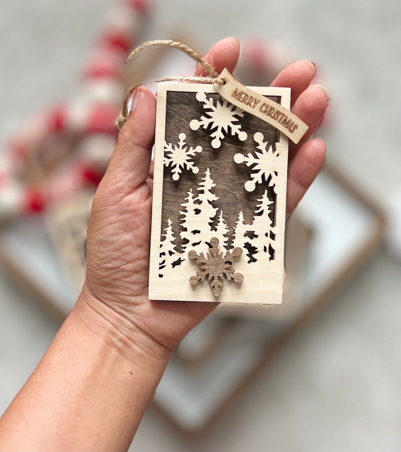May include: A wooden Christmas ornament with a cutout design of a forest and snowflakes. The ornament is brown and has a small wooden tag that says "Merry Christmas".