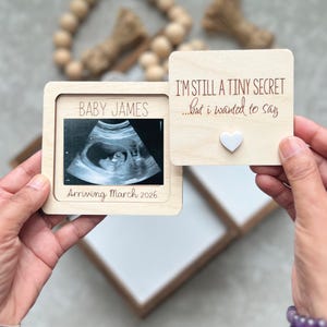 May include: A wooden pregnancy announcement frame with the text "BABY JAMES" and "Arriving March 2026". It holds an ultrasound image and a separate piece with the text "I'M STILL A TINY SECRET...but I wanted to say" and a heart.