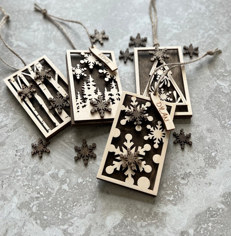 May include: Four wooden Christmas ornaments with different designs. Each ornament has a snowflake cutout and a personalized name tag. The names on the tags are Dylan, Kylie, and two others.