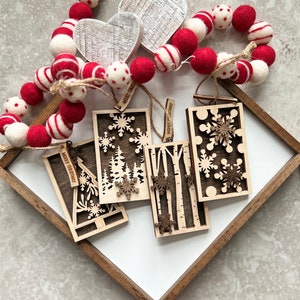 May include: Four wooden Christmas ornaments with laser cut designs of trees, snowflakes, and birch trees. Each ornament has a small tag with the text "Merry Christmas" on it. The ornaments are displayed on a white surface with a wooden frame.