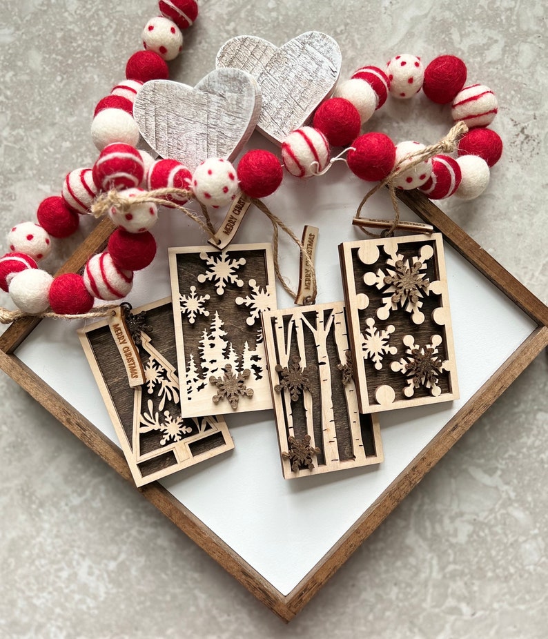 May include: Four wooden Christmas ornaments with laser cut designs of trees, snowflakes, and birch trees. Each ornament has a small tag with the text "Merry Christmas" on it. The ornaments are displayed on a white surface with a wooden frame.